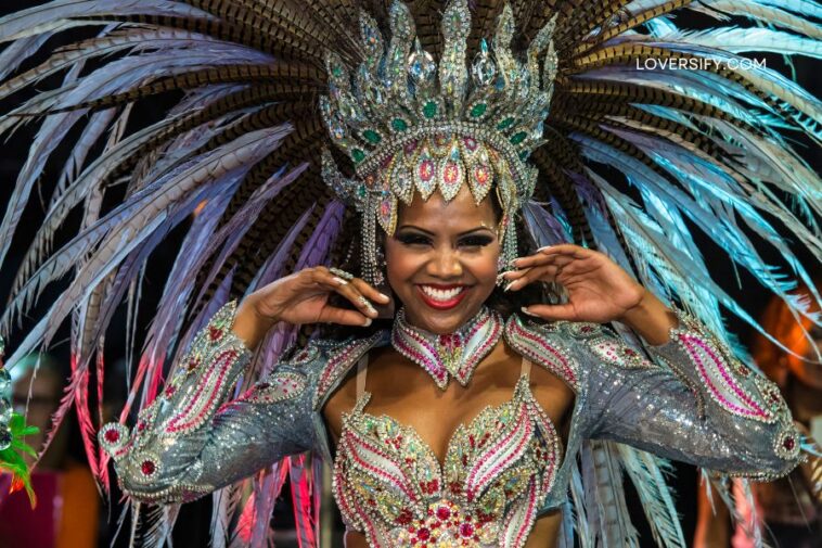 A smiling woman in a colorful, feathered carnival costume with elaborate headdress and sparkling embellishments.