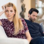 A woman with blonde hair rests her head on her hand while looking away from a man who is looking at his phone.