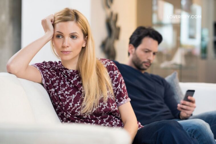A woman with blonde hair rests her head on her hand while looking away from a man who is looking at his phone.