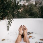 A person's feet, with manicured toenails, submerged in a white bath filled with milk and scattered flower petals, surrounded by lush greenery and large rocks.