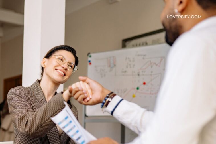 A smiling woman in a brown blazer shakes hands with a man in a white shirt, holding a document. A whiteboard with charts is visible in the background.