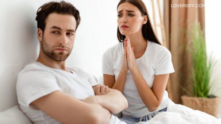 A man with arms crossed looks away from a woman in bed who clasps her hands together and looks at him pleadingly.
